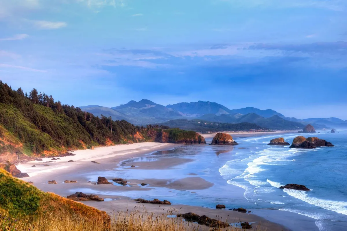 A striking view of the expansive sandy beach, rolling ocean waves, and dramatic rock formations, with distant mountains in the background. The golden hour lighting enhances the tranquil mood and showcases the broad natural beauty just outside the property.