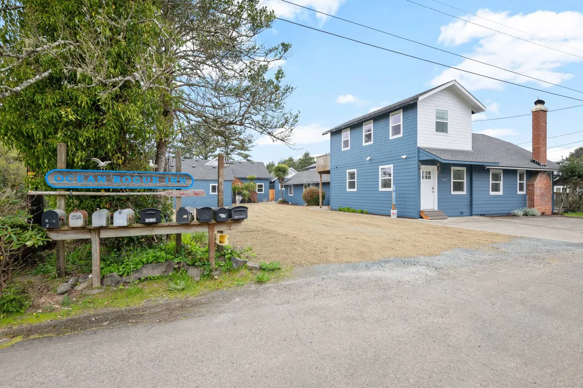 The blue exterior of the inn is shown along with a signboard reading 'Ocean Rogue Inn' and a row of mailboxes. The porch and front yard suggest a welcoming, social environment ideal for relaxing after a beach adventure.
