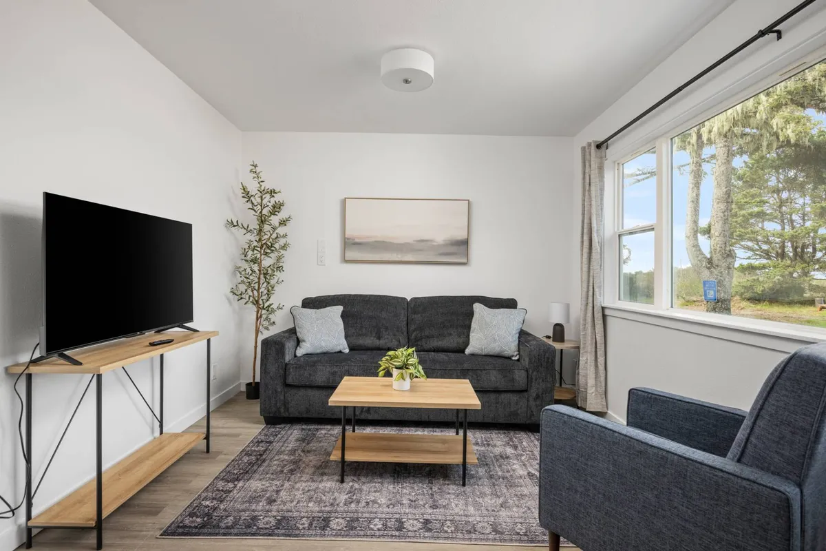 This cozy, light-filled living area features a plush sofa, stylish coffee table, smart TV, vibrant coastal artwork, and a lush potted plant, creating an inviting space perfect for unwinding after a day at the beach.