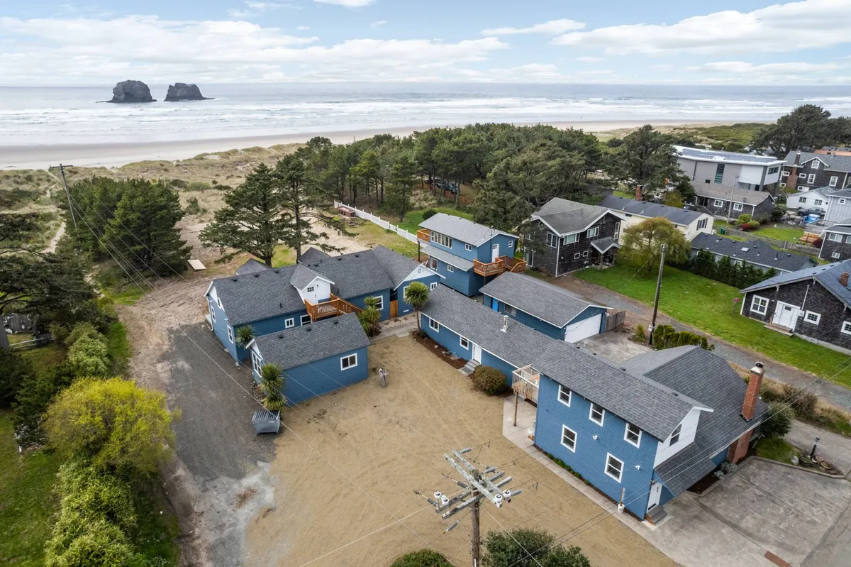 A wide exterior view of the beachfront property with nearby rock formations and surrounding trees, including an open courtyard area ideal for gatherings. The scene evokes a sense of adventure and provides context for proximity to the beach and nature.