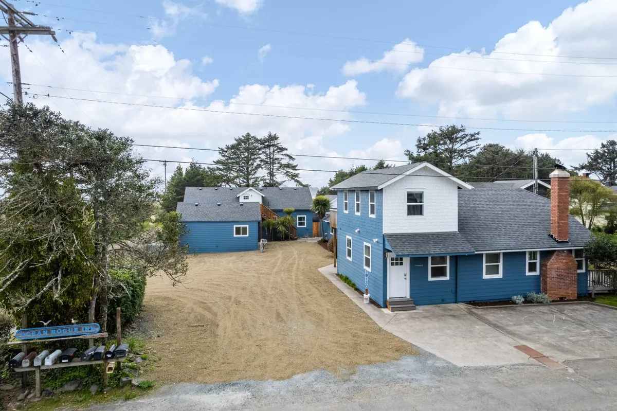 This image features the blue exterior of Ocean Rogue Inn with a red brick chimney, open yard space, and a visible signpost. The well-kept grassy yard and surrounding greenery provide plenty of outdoor space for both people and pets to explore and relax.
