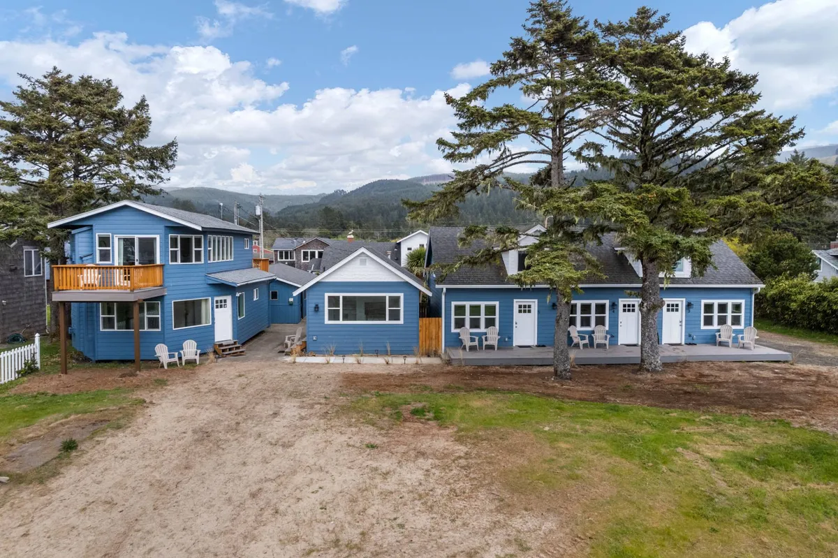 A cheerful exterior shot highlighting the blue house facade with a spacious deck and large trees, suggesting spaces where guests can relax outdoors, enjoy sunset views, or drink coffee in the morning. The inviting deck and lush surroundings evoke the natural beauty and laid-back atmosphere of the Oregon coast.