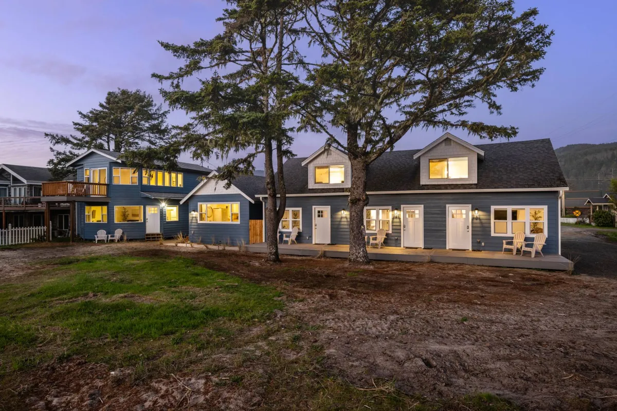 A welcoming two-story building sits beneath tall coastal trees, with its illuminated windows and a cozy front porch inviting guests to relax. People are gathered around an outdoor fire pit, while others enjoy the porch, hinting at both social and peaceful moments. The overall mood is peaceful and inviting, perfect for unwinding after a day at the beach.