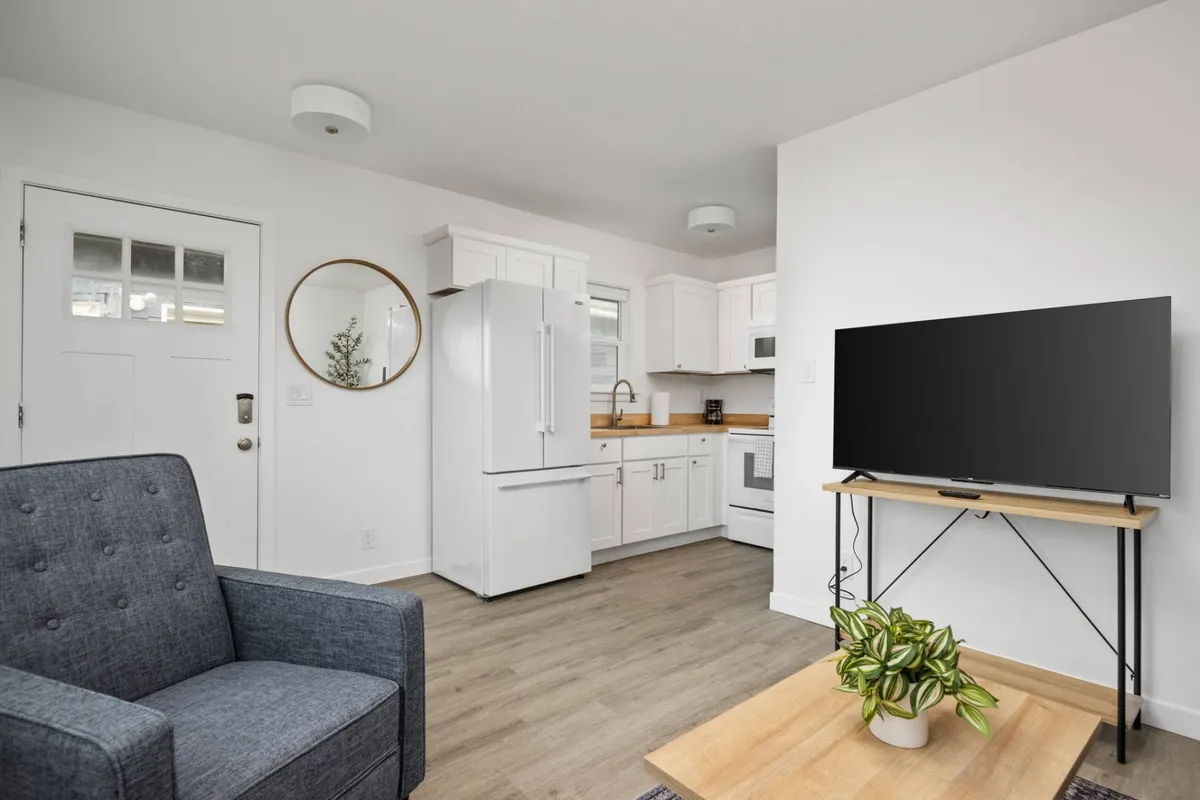 An open-concept living space featuring a plush sofa, a modern kitchen with dining table, a television, and contemporary décor. Ample natural light fills the room, and the arrangement allows for both lounging and socializing. The kitchen is fully equipped, though a dishwasher is not visible, and the overall aesthetic is clean, inviting, and modern-coastal.