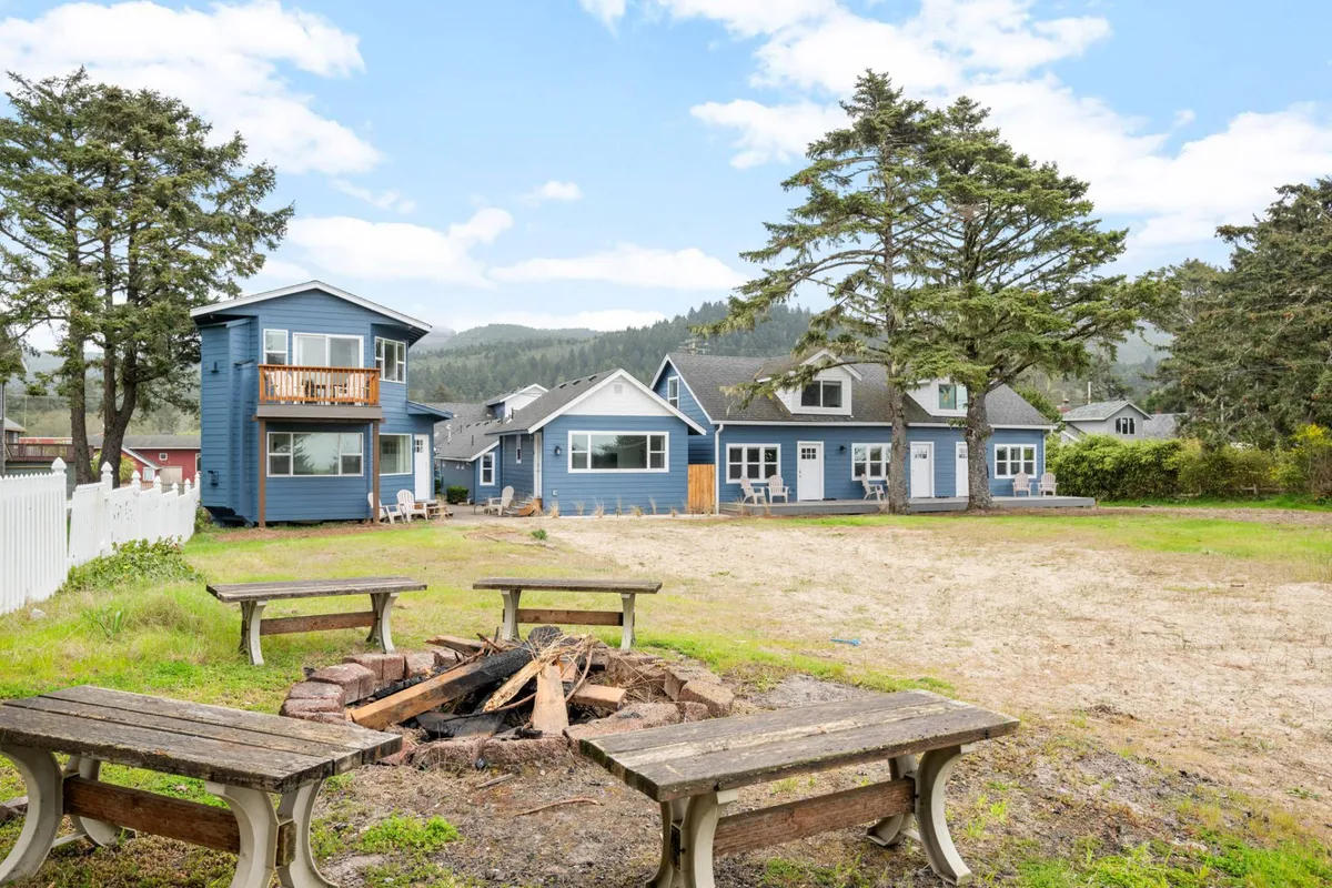 This image centers around a communal fire pit surrounded by blue cottages and large trees, exuding a relaxed and sociable atmosphere. The fire pit area features comfortable seating, inviting guests to gather for storytelling and camaraderie as dusk falls.